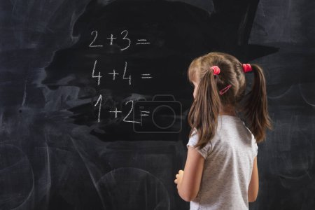 Photo for Schoolgirl standing in front of a blackboard, solving math equations, practicing summation - Royalty Free Image