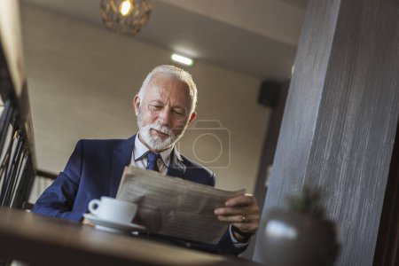 Photo for Senior businessman sitting in a modern office building restaurant, drinking coffee and reading newspapers - Royalty Free Image