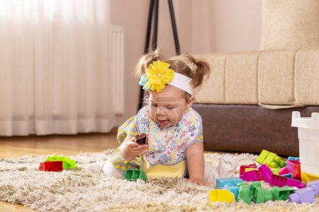 Foto de Hermosa niña, sentada en una alfombra en el piso del vivero, jugando con bloques de juguetes y sonriendo felizmente - Imagen libre de derechos