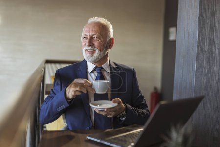 Foto de Hombre de negocios sénior sentado en una mesa de restaurante, tomando una taza de café y leyendo noticias en la computadora portátil - Imagen libre de derechos