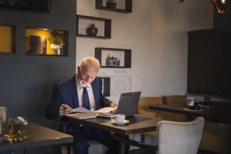 Foto de Hombre de negocios sénior en una oficina moderna trabajando en una computadora portátil y analizando contratos y documentación, seria y pensativa - Imagen libre de derechos