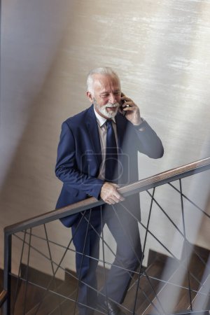 Téléchargez les photos : Homme d'affaires senior debout sur l'escalier moderne de l'immeuble de bureaux, ayant une conversation téléphonique - en image libre de droit