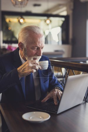 Téléchargez les photos : Homme d'affaires senior assis à une table de restaurant, prenant une tasse de café et lisant les nouvelles sur un ordinateur portable - en image libre de droit