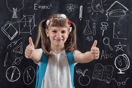 Photo for Schoolgirl wearing backpack, standing in front of blackboard and showing thumbs up on her first day of school - Royalty Free Image