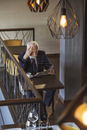 Foto de Vista de alto ángulo del hombre de negocios sénior sentado en un moderno restaurante edificio de oficinas, beber café y leer el periódico - Imagen libre de derechos