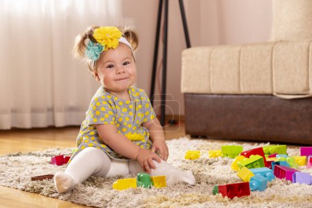 Téléchargez les photos : Belle petite fille, assise sur un tapis sur le sol de la pépinière, jouant avec des blocs de jouets et souriant joyeusement - en image libre de droit