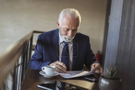 Téléchargez les photos : Homme d'affaires principal assis à une table de restaurant, analysant la documentation et les contrats et buvant du café - en image libre de droit