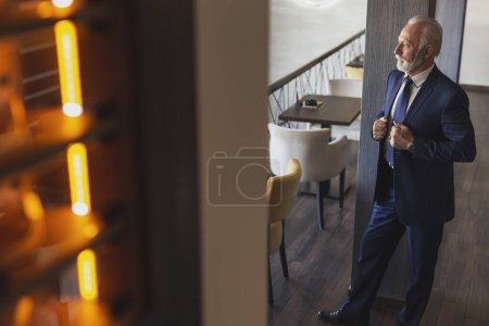 Foto de Retrato de un hombre de negocios pensativo y mayor de pie en un moderno restaurante de oficinas, apoyado en la pared y mirando a la distancia - Imagen libre de derechos