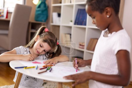 Foto de Dos niñas sentadas en el piso de una sala de juegos, dibujando con lápices de colores, terminando su proyecto de arte para la escuela - Imagen libre de derechos