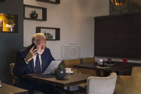 Téléchargez les photos : Homme d'affaires principal dans un restaurant, ayant une conversation téléphonique tout en travaillant sur une tablette - en image libre de droit