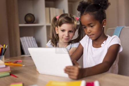 Téléchargez les photos : Deux petites filles assises à un bureau, jouant à des jeux vidéo sur une tablette et s'amusant. Concentrez-vous sur la fille à gauche - en image libre de droit