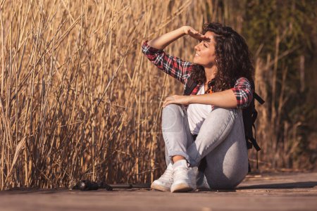 Téléchargez les photos : Femme assise sur les quais du lac en bois, prenant une pause de randonnée dans la nature, se détendre et profiter d'une journée d'automne ensoleillée - en image libre de droit
