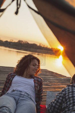 Foto de Pareja joven enamorada acampando en los muelles del lago, acostada en la entrada de la tienda, disfrutando de una hermosa puesta de sol y leyendo el mapa - Imagen libre de derechos