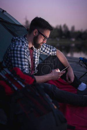 Photo for Young man camping on the lake docks, sitting on the tent entrance and typing a text message - Royalty Free Image
