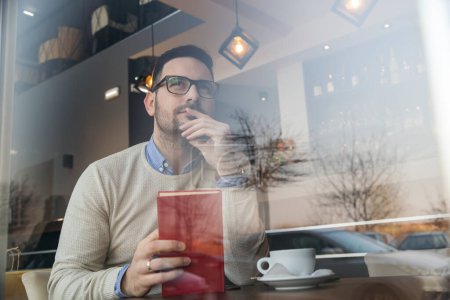 Hombre guapo sentado en una mesa de restaurante, bebiendo café y sosteniendo un libro, mirando pensativamente la distancia, perdido en el pensamiento