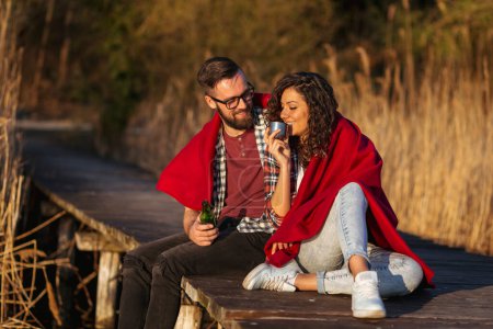 Téléchargez les photos : Couple assis sur les quais du lac, couvert de couverture, boire de la bière et du punch chaud et s'amuser - en image libre de droit