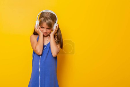 Téléchargez les photos : Portrait de belle petite fille vêtue d'une robe bleue et d'un casque, écoutant la musique les yeux fermés, isolée sur fond jaune - en image libre de droit