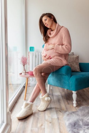 Foto de Hermosa joven embarazada relajándose en casa y esperando a su bebé; mujer embarazada sentada en un sillón, sosteniendo su vientre y sonriendo - Imagen libre de derechos