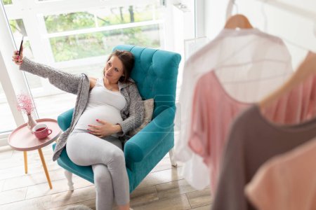 Foto de Mujer embarazada relajándose en casa, sentada en un sillón, bebiendo té y tomando selfies con un smartphone - Imagen libre de derechos