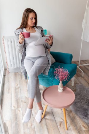 Photo for Beautiful pregnant woman relaxing at home, sitting in an armchair and reading maternity blogs on a tablet computer - Royalty Free Image