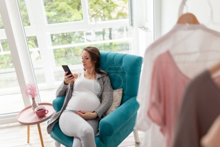 Téléchargez les photos : Femme enceinte assise dans un fauteuil, relaxante à la maison, buvant une tasse de thé chaud et utilisant un téléphone intelligent - en image libre de droit