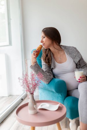 Photo for Beautiful pregnant woman sitting in an armchair, drinking glass of milk and eating croissant for breakfast - Royalty Free Image