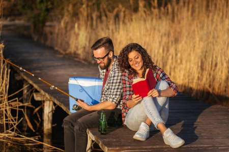 Téléchargez les photos : Couple assis sur les quais du lac, l'homme pêche pendant que la femme lit un livre, profiter d'une journée paisible dans la nature et se détendre - en image libre de droit
