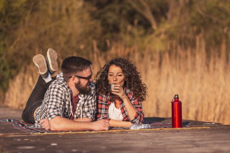 Foto de Hermosa pareja joven enamorada de hacer un picnic en los muelles del lago, acostada en una manta de picnic, tomando café y disfrutando de un hermoso día soleado de otoño en la naturaleza - Imagen libre de derechos