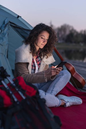 Photo for Young woman camping by the lake, sitting near the tent entrance and surfing the Internet using smart phone - Royalty Free Image