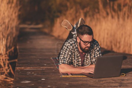 Foto de Hombre acostado en los muelles del lago, usando una computadora portátil, escribiendo una entrada de blog, trabajando remotamente mientras se relaja en la naturaleza; freelancer masculino trabajando al aire libre, disfrutando de un día soleado de otoño - Imagen libre de derechos