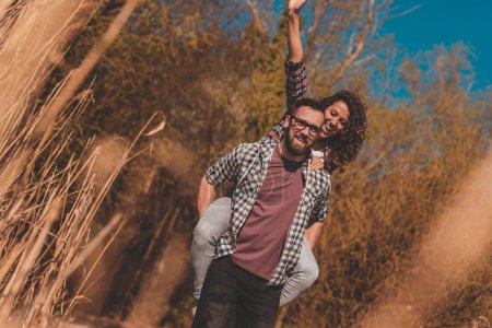 Téléchargez les photos : Couple en amour avoir du plaisir en plein air, homme piggybackking femme tout en marchant sur les quais du lac et rire - en image libre de droit