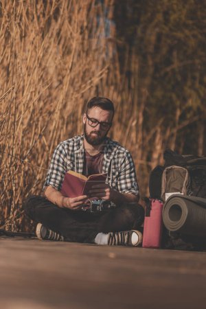 Téléchargez les photos : Homme assis sur les quais du lac en bois, profitant d'une belle journée d'automne dans la nature et lisant un livre - en image libre de droit