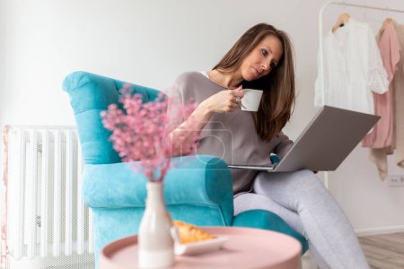 Photo for Beautiful pregnant woman sitting in an armchair, using a laptop computer and drinking coffee - Royalty Free Image