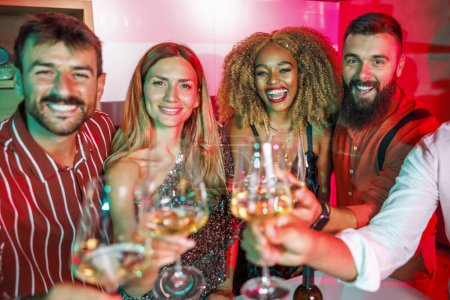 Photo for Group of cheerful young friends having fun at house party, making a toast raising glasses of wine towards the camera, dancing and drinking - Royalty Free Image