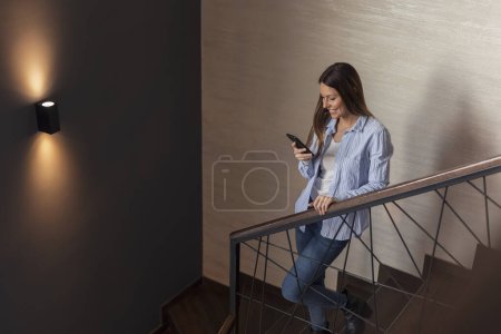 Téléchargez les photos : Belle jeune femme debout sur un escalier, souriant en tapant un message texte sur un téléphone intelligent - en image libre de droit