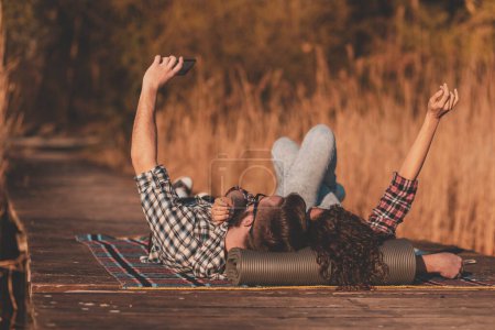 Foto de Hermosa pareja enamorada acostada en una manta de picninc en los muelles del lago, tomando una selfie y divirtiéndose en un día soleado de otoño - Imagen libre de derechos