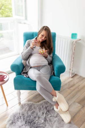 Foto de Retrato de una hermosa mujer embarazada sentada en un sillón, comiendo ensalada mixta de verduras de temporada; concepto de embarazo y dieta saludable - Imagen libre de derechos