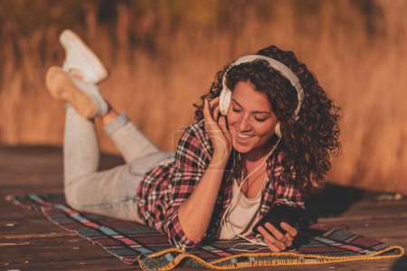 Téléchargez les photos : Femme portant un casque couché sur les quais du lac, utilisant un téléphone intelligent, choisissant une chanson dans une playlist, écoutant la musique et chantant un jour ensoleillé d'automne - en image libre de droit