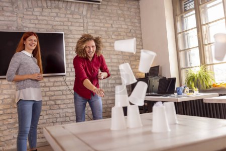 Foto de Gente de negocios divirtiéndose en la oficina, jugando juegos de oficina mientras están en un descanso, derribando una pirámide de vasos de plástico con una bola de papel - Imagen libre de derechos