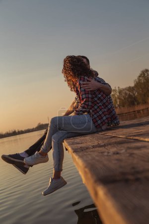Photo for Beautiful young couple in love sitting at lake docks, hugging and enjoying a beautiful sunset over the lake - Royalty Free Image