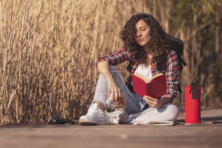 Téléchargez les photos : Belle femme assise sur les quais en bois, profitant d'une journée d'automne dans la nature, lisant un livre et mangeant un sandwich au bord du lac - en image libre de droit
