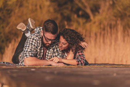 Téléchargez les photos : Couple amoureux couché sur une couverture de pique-nique sur les quais du lac, s'amuser et profiter d'une belle journée ensoleillée d'automne - en image libre de droit