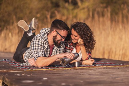 Téléchargez les photos : Jeune couple pique-niquer sur les quais du lac, verser du café dans une bouteille thermos et profiter de la journée ensoleillée d'automne en plein air - en image libre de droit