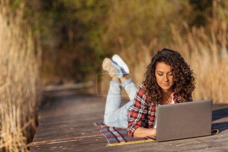 Téléchargez les photos : Femme pigiste couchée sur les quais du lac, travaillant sur un ordinateur portable tout en se relaxant dans la nature - en image libre de droit