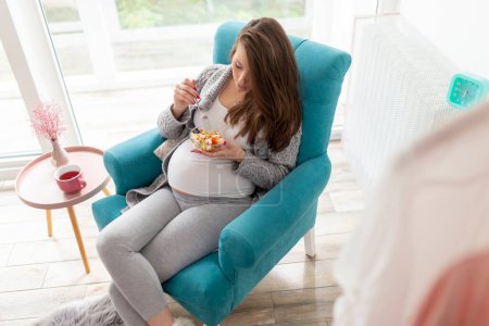 Téléchargez les photos : Belle femme enceinte assise dans un fauteuil, mangeant une salade mixte de légumes de saison ; concept de grossesse et d'alimentation saine - en image libre de droit