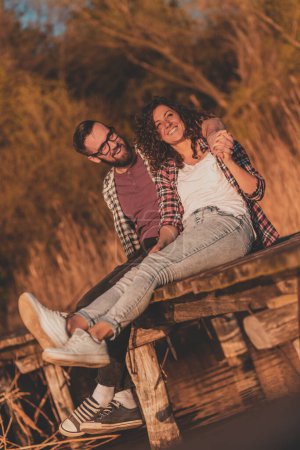 Photo for Beautiful young couple in love sitting at lake docks, hugging and enjoying a beautiful sunset over the lake - Royalty Free Image