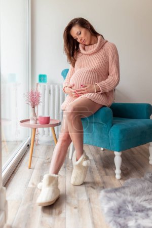 Photo for Beautiful young pregnant woman relaxing at home and expecting her baby; pregnant woman sitting in an armchair, holding and cuddling her belly - Royalty Free Image