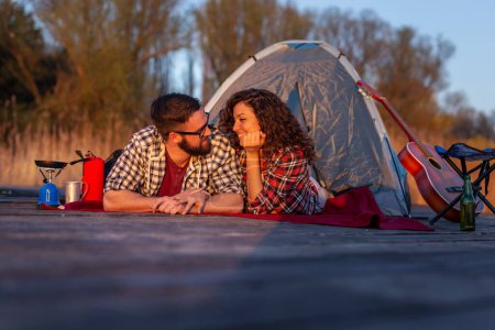 Photo for Couple in love camping at the lake docks, lying at the tent entrance and enjoying beautiful sunset - Royalty Free Image