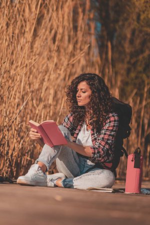 Téléchargez les photos : Étudiante assise sur les quais du lac en bois, profitant d'une belle journée d'automne dans la nature et étudiant pour une période d'examen - en image libre de droit