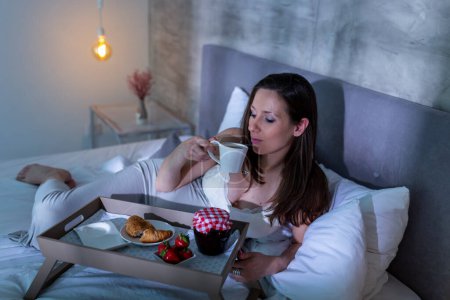 Foto de Hermosa mujer embarazada con un camisón acostado en la cama al lado de una cena servida en una bandeja, bebiendo té; mujer embarazada y concepto de nutrición saludable - Imagen libre de derechos